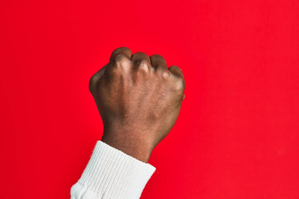 Χέρι και χέρι του αφροαμερικανού μαύρου άνδρα πάνω από το κόκκινο απομονωμένο υπόβαθρο κάνει διαμαρτυρία και χειρονομία επανάστασης, γροθιά εκφράζοντας δύναμη και δύναμη  - Φωτογραφία, εικόνα