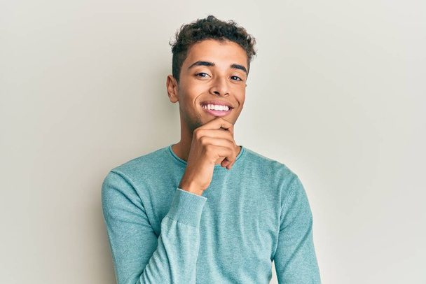Jonge knappe Afro-Amerikaanse man in casual kleding glimlachend zelfverzekerd kijkend naar de camera met gekruiste armen en hand op kin. positief denken.  - Foto, afbeelding