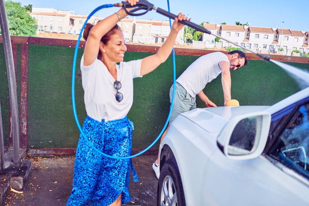 Middle age beautiful couple wearing casual clothes and smiling happy. Standing with smile on face washing car using water pistol and sponge. - Foto, immagini