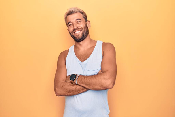 Jonge knappe blonde man met casual mouwloos t-shirt over geïsoleerde gele achtergrond vrolijk gezicht glimlachend met gekruiste armen kijkend naar de camera. Positief persoon. - Foto, afbeelding