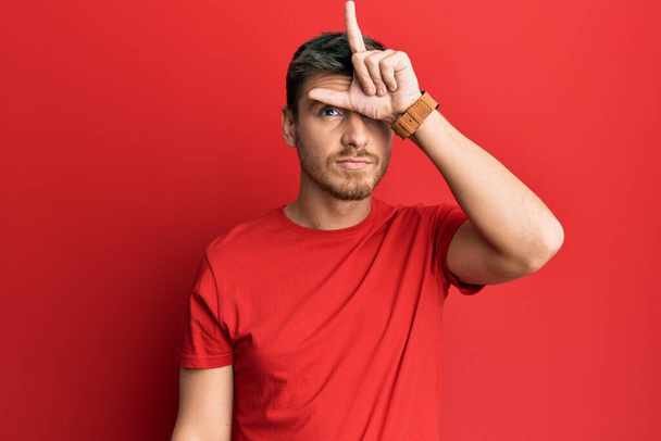 Handsome caucasian man wearing casual red tshirt making fun of people with fingers on forehead doing loser gesture mocking and insulting.  - Photo, Image