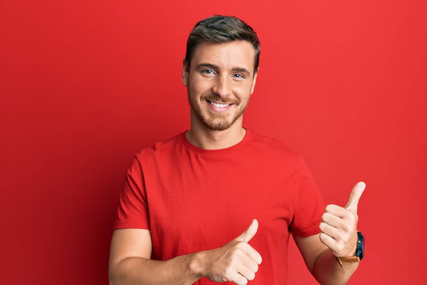 Красивый белый мужчина в обычной красной футболке, указывающей на спину с поднятой рукой и большим пальцем, уверенно улыбающийся  - Фото, изображение
