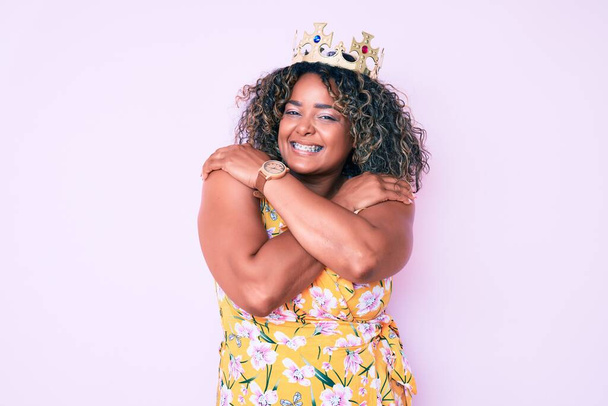 Young african american plus size woman wearing princess crown hugging oneself happy and positive, smiling confident. self love and self care  - Foto, Imagen