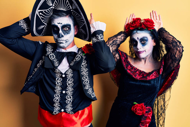 Ein junges Paar im mexikanischen Totenkostüm über gelben Hasenohren gestikuliert mit Handflächen, die zynisch und skeptisch aussehen. Osterhasen-Konzept.  - Foto, Bild