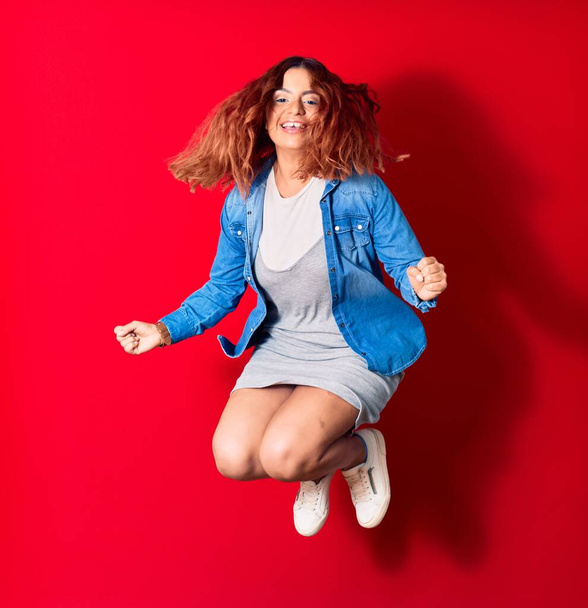 Young beautiful latin woman wearing casual clothes smiling happy. Jumping with smile on face doing winner gesture with fists up over isolated red background - Photo, Image