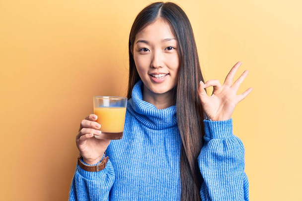 Młoda piękna Chinka pije szklankę soku pomarańczowego robi ok znak palcami, uśmiechając przyjazny gest doskonały symbol  - Zdjęcie, obraz