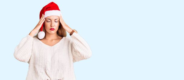 Piękna młoda kobieta nosząca świąteczny kapelusz cierpiąca na ból głowy zdesperowana i zestresowana, ponieważ ból i migrena. ręce na głowę.  - Zdjęcie, obraz