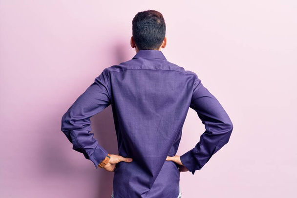 Νεαρός όμορφος άντρας με γενειάδα φορώντας ένα απλό πουκάμισο να στέκεται προς τα πίσω κοιτάζοντας μακριά με τα χέρια στο σώμα  - Φωτογραφία, εικόνα