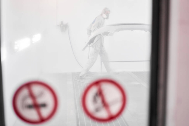 All in a haze of paint. Worker painting parts of the car in special painting chamber, wearing costume and protective gear. Car service station. - Photo, Image