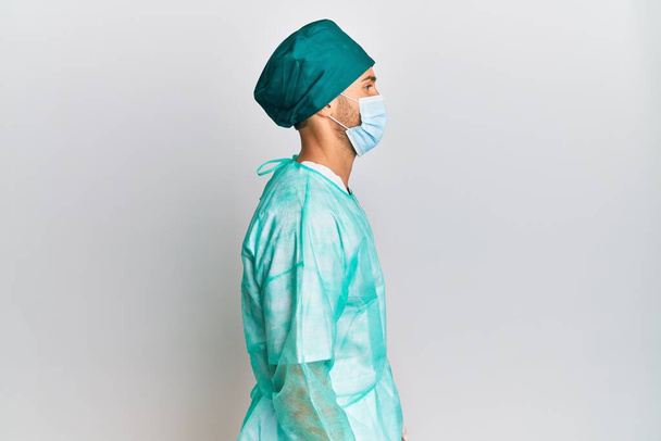 Young handsome man wearing surgeon uniform and medical mask looking to side, relax profile pose with natural face with confident smile.  - Photo, Image