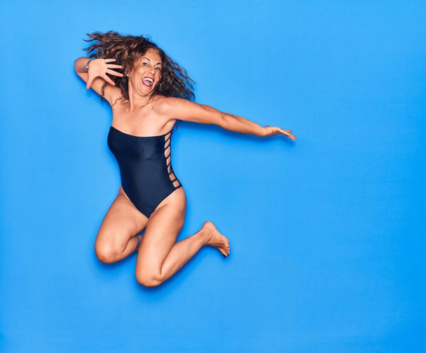 Middle age beautiful hispanic woman on vacation wearing swimsuit smiling happy. Jumping with smile on face over isolated blue background - Фото, зображення