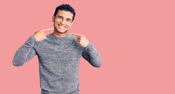 Latino komea nuori mies yllään rento villapaita hymyilevä iloinen osoittaa ja osoittaa sormilla hampaat ja suu. hammasterveyden käsite.  - Valokuva, kuva