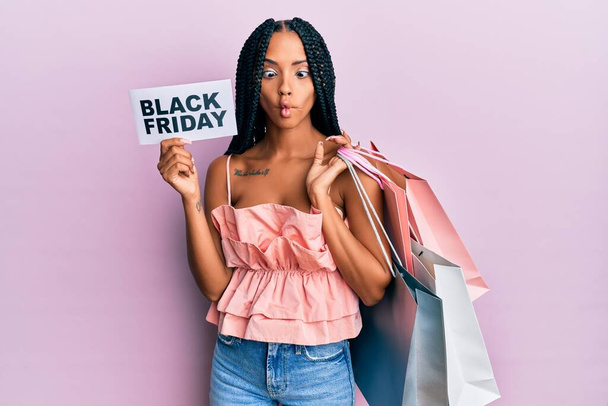 Beautiful hispanic woman holding shopping bags and black friday banner making fish face with mouth and squinting eyes, crazy and comical.  - Fotó, kép