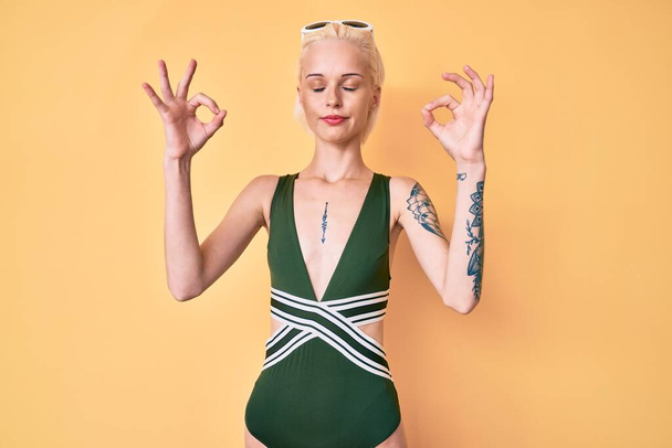 Jonge blonde vrouw met tatoeage in badpak en zonnebril ontspannen en glimlachend met gesloten ogen meditatiegebaar doen met vingers. yoga concept.  - Foto, afbeelding