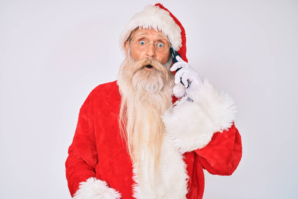 Старик в костюме Санта-Клауса, говорит по телефону испуганный и изумленный открытыми ртами в ожидании сюрприза, неверного лица  - Фото, изображение