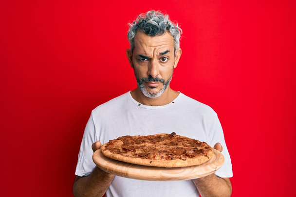 Grauhaariger Mann mittleren Alters, der eine italienische Pizza in der Hand hält, skeptisch und nervös, der wegen des Problems die Stirn runzelt. Negative Person.  - Foto, Bild