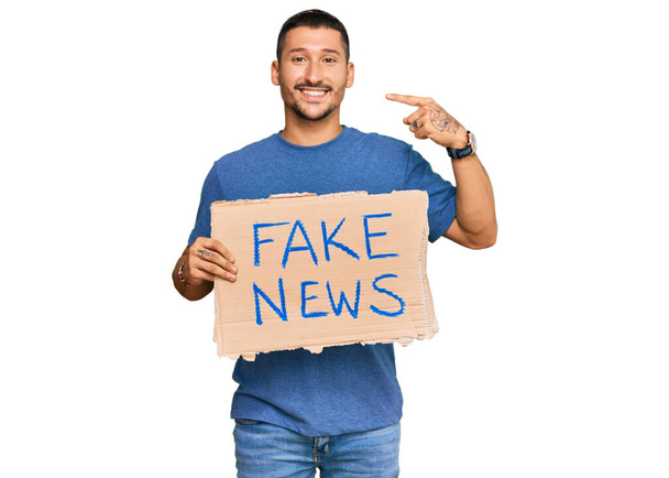 Hombre guapo con tatuajes sosteniendo banner de noticias falsas apuntando con el dedo a uno mismo sonriendo feliz y orgulloso  - Foto, Imagen