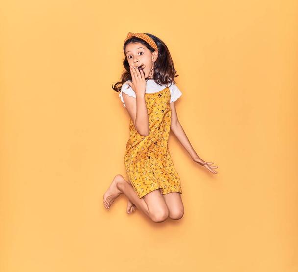Roztomilé hispánské dítě dívka na sobě ležérní šaty a diadém s úsměvem šťastný. Skákání s úsměvem na tváři přes izolované žluté pozadí - Fotografie, Obrázek
