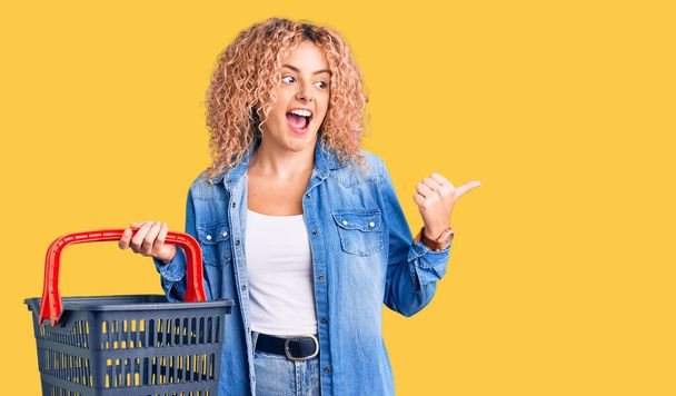 Joven mujer rubia con el pelo rizado sosteniendo cesta de la compra del supermercado apuntando el pulgar hacia el lado sonriendo feliz con la boca abierta  - Foto, imagen