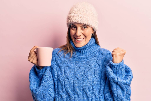 Kışlık giysiler giymiş genç bir kadın elinde kahveyle gururla bağırıyor zaferi kutluyor ve başarıyı kollarını kaldırarak kutluyor.  - Fotoğraf, Görsel