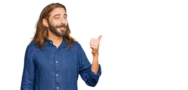 Atrakcyjny mężczyzna z długimi włosami i brodą w luźnych ubraniach uśmiechnięty ze szczęśliwym wyglądem twarzy i skierowany na bok z kciukiem w górze.  - Zdjęcie, obraz