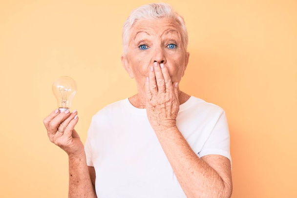 Senior beautiful woman with blue eyes and grey hair holding lightbulb for inspiration and idea covering mouth with hand, shocked and afraid for mistake. surprised expression  - Photo, Image