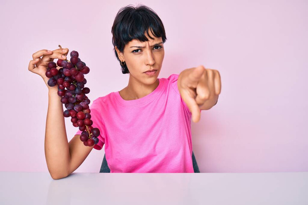 Hermosa morena mujer sosteniendo rama de uvas frescas señalando con el dedo a la cámara y a usted, gesto de confianza mirando en serio  - Foto, imagen