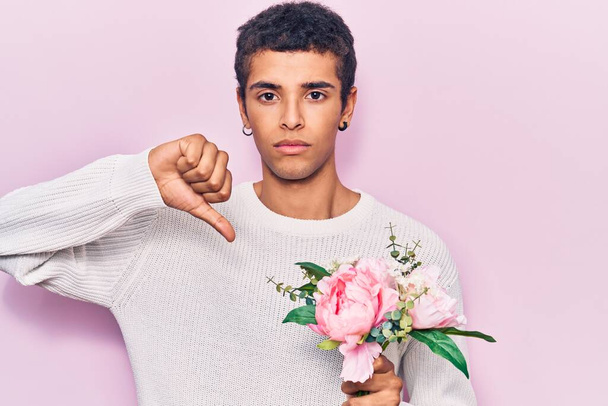 怒った顔の花を持つ若いアフリカ系アメリカ人男性、親指で嫌いであることを示す負の兆候、拒絶概念  - 写真・画像