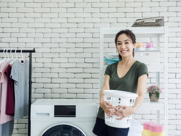 Mooie jonge Aziatische vrouw huisvrouw staande en het houden van lege witte doek mand met glimlachen in de buurt van wasmachine in wasruimte. - Foto, afbeelding