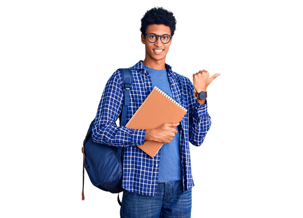 Jovem afro-americano usando mochila estudantil segurando livro apontando para as costas com a mão e os polegares para cima, sorrindo confiante  - Foto, Imagem