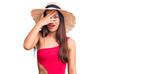 Fiatal gyönyörű latin lány fürdőruhát visel és nyári kalap kukucskál sokk borító arc és a szemek kézzel, néz keresztül ujjak zavarba ejtő kifejezés.  - Fotó, kép