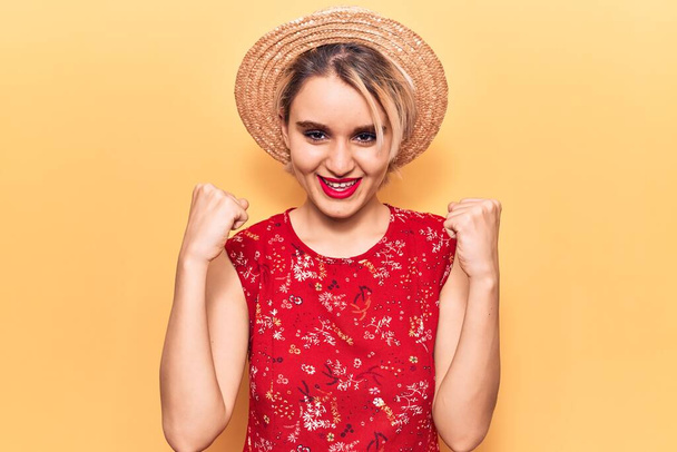 Jovem mulher loira bonita usando chapéu de verão gritando orgulhoso, celebrando vitória e sucesso muito animado com braços levantados  - Foto, Imagem