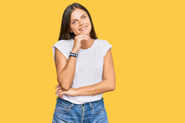 Young caucasian woman wearing casual white tshirt looking confident at the camera with smile with crossed arms and hand raised on chin. thinking positive.  - Photo, Image