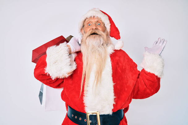 Alter älterer Mann mit grauen Haaren und langem Bart im Weihnachtsmann-Kostüm mit Einkaufstüten, die den Sieg mit glücklichem Lächeln und Siegerausdruck mit erhobenen Händen feiern  - Foto, Bild