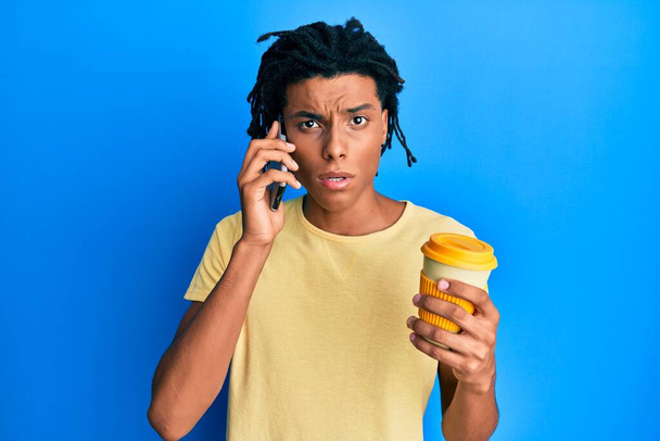 Jeune homme afro-américain utilisant un smartphone et buvant une tasse de café dans le visage de choc, l'air sceptique et sarcastique, surpris avec la bouche ouverte  - Photo, image
