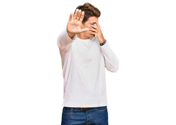 Hombre caucásico guapo vistiendo suéter blanco casual cubriendo los ojos con las manos y haciendo gesto de parada con expresión triste y de miedo. concepto avergonzado y negativo.  - Foto, Imagen