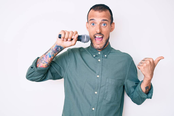 Młody przystojny mężczyzna z tatuażem śpiewający piosenkę za pomocą mikrofonu skierowanego kciukiem w górę do boku uśmiechnięty szczęśliwy z otwartymi ustami  - Zdjęcie, obraz