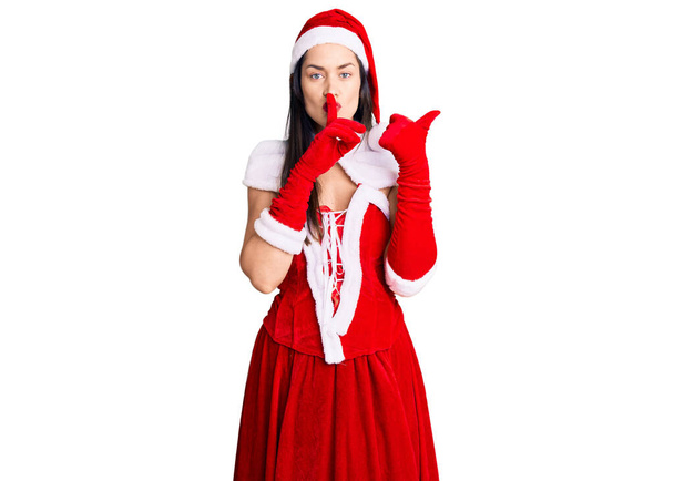 Νεαρή όμορφη καυκάσια γυναίκα που φοράει κοστούμι Santa Claus ζητώντας να είναι ήσυχο με το δάχτυλο στα χείλη δείχνοντας με το χέρι στο πλάι. σιωπή και μυστική έννοια.  - Φωτογραφία, εικόνα