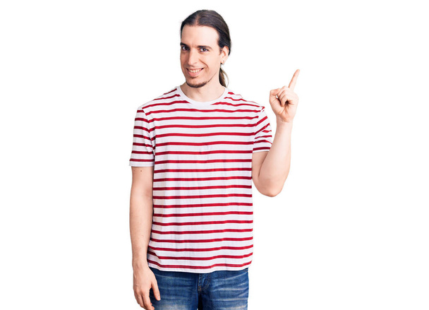 Fiatal felnőtt férfi hosszú haj visel alkalmi csíkos póló mosolygós boldog mutató kéz és ujj oldalra  - Fotó, kép