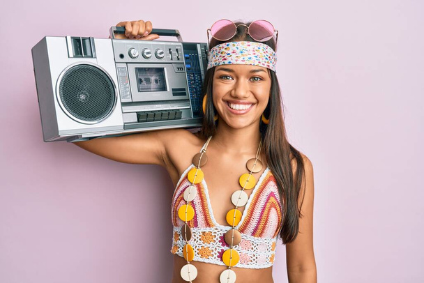 Hermosa mujer hispana vistiendo estilo hippie sosteniendo la caja de boom con un aspecto positivo y feliz de pie y sonriendo con una sonrisa confiada mostrando los dientes  - Foto, Imagen