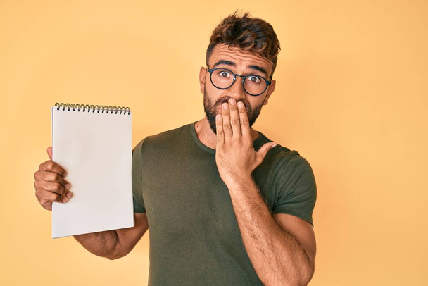 Jonge Spaanse man draagt een bril met notitieboekje over mond met hand, geschokt en bang voor fouten. verraste uitdrukking  - Foto, afbeelding