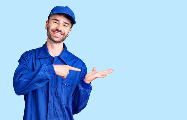 Mladý hispánec v malířské uniformě užaslý a usmívající se do kamery při prezentaci rukou a ukazující prstem.  - Fotografie, Obrázek