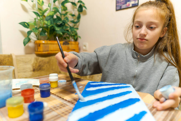 девочка рисует голубую картонку из гуаши, делает фон, сидит на домашней кухне - Фото, изображение
