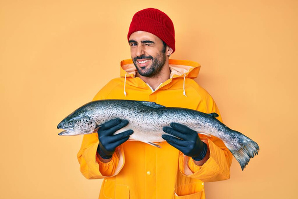 Yakışıklı, sakallı, balıkçı kıyafeti giyen yakışıklı bir adam kameraya seksi bir ifadeyle bakıyor, neşeli ve mutlu bir yüzle..  - Fotoğraf, Görsel