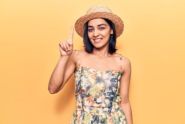 Young beautiful latin woman wearing summer hat showing and pointing up with finger number one while smiling confident and happy.  - Photo, Image
