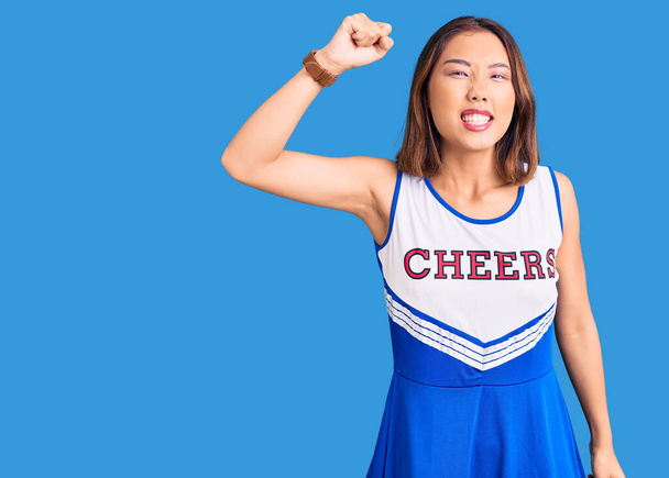 Giovane bella ragazza cinese indossa uniforme cheerleader arrabbiato e pazzo alzando il pugno frustrato e furioso, mentre gridando con rabbia. rabbia e concetto aggressivo.  - Foto, immagini