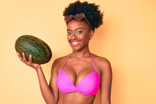 Giovane donna afroamericana in bikini con in mano melone dall'aspetto positivo e felice e sorridente con un sorriso fiducioso che mostra i denti  - Foto, immagini