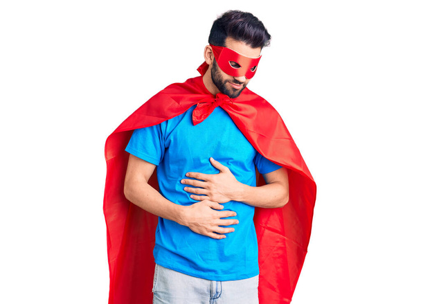 Junger gutaussehender Mann mit Bart im Superheldenkostüm mit der Hand auf dem Bauch wegen Verdauungsstörungen, schmerzhafter Krankheit, die sich unwohl anfühlt. Schmerzkonzept.  - Foto, Bild