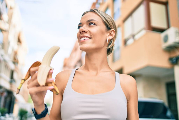 Νεαρή καυκασιάτισσα γυμνάστρια που φοράει αθλητικά ρούχα προπονείται σε εξωτερικούς χώρους τρώγοντας υγιή μπανάνα για δύναμη και ενέργεια - Φωτογραφία, εικόνα