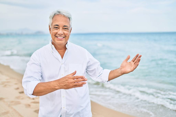 Handosme hispánský muž s šedivými vlasy se šťastně usmívá na pláži, těší dovolená ukazuje na kopírovací prostor - Fotografie, Obrázek